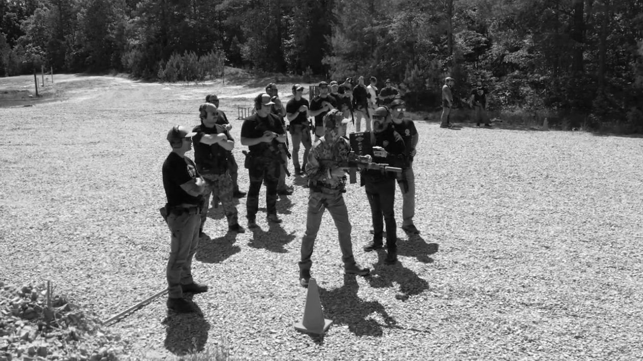 A DNC Defense class gathered on a gravel range, instructor demonstrating with a carbine
